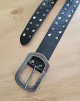 Cinturón de cuero negro con textura granulada, decorado con tachas metálicas redondas en tonos plateados y bronce. Posee una hebilla metálica grande de estilo envejecido con forma rectangular redondeada y un pasante metálico.