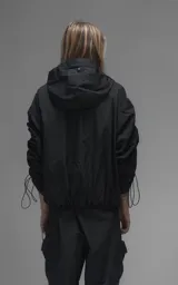 Campera rompevientos negra de calce oversize, confeccionada en microfibra técnica ligera con acabado semi-mate. Presenta capucha integrada, cierre frontal técnico y bolsillos tipo ojal. Destaca por su sistema de frunces elásticos regulables mediante tancas a lo largo de las mangas y el ruedo, permitiendo ajustar el volumen de la prenda.