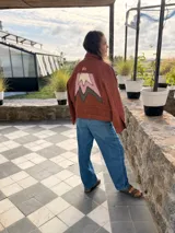Campera bomber de jean color terracota, con corte holgado. Presenta apliques de cuero cortados y bordados a mano en la espalda, delantera y cuello, formando un diseño de montañas en tonos beige, rosa y verde militar.