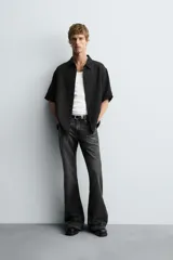 Camisa negra de manga corta con cuello solapa y textura arrugada o plisada. Corte relajado (relaxed fit) con aberturas laterales y cierre frontal de botones.