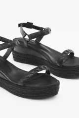 Sandalia negra con plataforma forrada en yute trenzado, de doble tira. La tira de la capellada está confeccionada en cuero con tachas texturadas y lisas de acabado níquel, mientras que el tobillo se sujeta con una segunda tira de cuero con las mismas tachas y hebilla metálica con grabada de la marca. Cuenta con una suela de caucho para mayor confort.