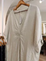 Vestido midi de lino color beige, con diseño oversize, escote en V y detalle de nudo drapeado en el frente. Posee mangas cortas amplias con dobladillo y corte asimétrico, siendo más largo en la parte trasera.