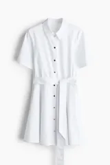 Vestido camisero corto color blanco, de manga corta, con botones metálicos frontales, cuello camisero, falda circular y cinturón de la misma tela para anudar en la cintura.