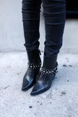 Bota texana corta de cuero negro con textura de piel de serpiente, punta fina y taco cubano. Presenta arnés desmontable con apliques metálicos plateados.