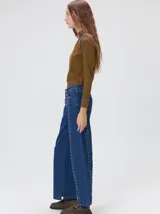 Jeans de corte ancho (wide leg) y tiro medio, confeccionados en denim de algodón negro con costuras contrastantes en color ocre. Presenta múltiples botones metálicos frontales y tachas metálicas aplicadas a lo largo de los laterales y la pretina.