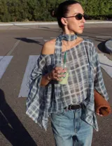 Blusa fluida y amplia con estampa a cuadros en tonos azules y blancos, diseño asimétrico con hombro descubierto, mangas amplias y tira larga integrada al cuello.