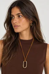 Collar largo con cordón de cuero tostado y dije metálico ovalado en tono dorado envejecido.