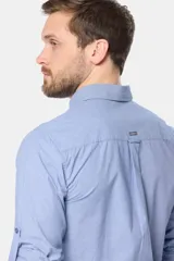 Camisa celeste de manga larga confeccionada en algodón, con cuello camisero, cierre frontal de botones, bolsillo en el pecho y diseño de cuadros sutiles.