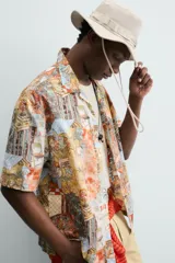 Camisa de corte relajado confeccionada en tejido ligero de algodón, con cuello bowling y manga corta. Presenta un estampado integral con motivos urbanos y tropicales en tonos cálidos.