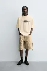 Camiseta de algodón de corte relajado, color beige claro, con cuello redondo y manga corta. Presenta un estampado central de garabatos negros con texto en mayúsculas que dice "TAKE RIGHT STEP FIND A RIGHT CORD".