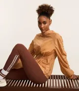 Campera deportiva femenina tipo parka, color ocre, confeccionada en poliamida y poliéster. Tiene cuello con capucha, mangas largas con puños elásticos, cierre frontal con cremallera y cordón ajustable en la cintura.