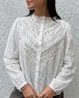 Camisa blanca de estilo romántico con detalles de encaje y alforzas en el frente. Presenta cuello alto con volado, mangas largas con puños elásticos y cierre frontal de botones.