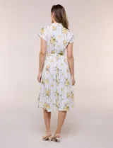 Vestido camisero midi de silueta evasé, confeccionado en tejido liviano blanco con estampado floral amarillo y rayas celestes. Cuenta con cuello solapa en V, cartera con botones frontales, mangas cortas japonesas y cintura definida con lazo ajustable a juego.