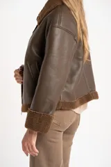 Campera tipo gamulán de cuero sintético marrón con interior y detalles de corderito en cuello, puños y ruedo. Presenta cierre frontal metálico y corte oversize.