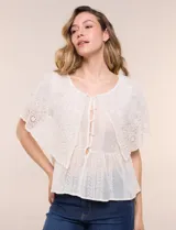 Blusa de estilo romántico color beige confeccionada en algodón, con escote amplio, importante volante capa en broderie calado, cartera frontal con botones tipo perla, bordados verticales calados, cintura marcada con bajo peplum y ruedo recto festonado.