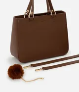 Cartera estilo satchel de tamaño mediano, confeccionada en poliuretano color marrón con textura tipo cuero. Cuenta con asas de mano reforzadas, correa larga desmontable, cierre superior de cremallera y un colgante decorativo de pompón de pelo sintético.