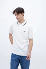 Remera polo blanca con cuello y puños acanalados con rayas azules y rojas. Presenta una media cartera de botones y un bordado en el pecho con la palabra "Uniform" en azul.