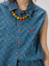 Chaleco clásico de calce cuadrado en color azul, con estampado geométrico de arcos entrelazados, pespuntes rojos en contraste, cuello camisero, cierre frontal de botones y gráfico bordado en el pecho.