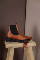 Bota tipo Chelsea de cuero marrón con suela track negra de goma. Presenta paneles elásticos laterales y una costura reforzada en la puntera. Incluye una correa de cuero desmontable con hebilla metálica.