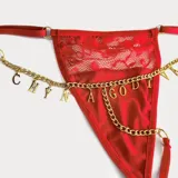 Tanga roja con diseño combinado de encaje floral en la parte superior y satén en la inferior, con tiras regulables y cadena metálica dorada con dijes de letras colgantes.