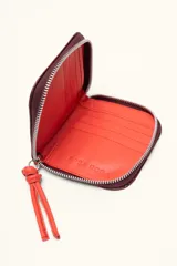 Billetera pequeña de cuero con textura tipo cocodrilo en color bordó, con cierre perimetral y detalle de tirador con nudo en color rojo.
