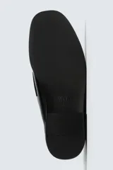 Mocasín de vestir negro con efecto acharolado, parte superior lisa con detalle de borlas, horma cuadrada y suela con ligero tacón.