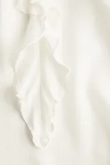 Blusa de crepé de gasa color blanco, con cuello mao, abertura frontal con tiras para anudar y detalle de volados verticales. Presenta mangas largas tipo globo de corte raglán con puños elásticos y terminación en volados.