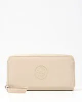 Billetera rectangular Extra Large de símil cuero color taupe, con cierre perimetral y tira corta para la muñeca. Presenta un aplique circular con el logo de la marca grabado en el centro.