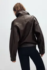 Cazadora tipo biker de cuero sintético en color marrón oscuro, con cuello subido, cierre frontal cruzado con botones forrados y cremallera oculta. Presenta mangas largas con trabillas en los hombros, bolsillos laterales de vivo y bajo con acabado elástico.