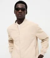 Campera tipo bomber masculina de color beige, con cuello, puños y ruedo acanalados. Presenta cierre frontal por cremallera, bolsillos laterales y acabado con tecnología repelente al agua.