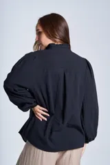 Camisa de color azul con cuello alto decorado con volados y lazo de ajuste. Presenta tablas decorativas en los hombros, cierre frontal con botones de nácar y mangas largas con puños elásticos que generan un efecto abullonado.