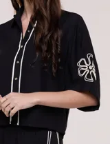 Conjunto de dos piezas negro compuesto por camisa de manga corta con cuello camisero y pantalón palazzo de tiro alto. Presenta detalles de bordados florales en contraste a lo largo de los laterales del pantalón y en las mangas.