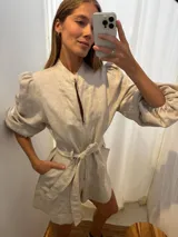 Mono corto de lino color beige, con cuello camisero, cierre frontal, mangas largas abullonadas y cinto ajustable en la cintura.
