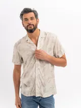 Camisa de manga corta color beige con estampado tropical de hojas, cuello camisero abierto y cierre frontal de botones.