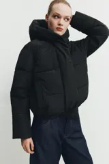 Campera puffer acolchada de color negro, con cuello envolvente, capucha integrada, manga larga, bolsillos laterales ocultos y bajo con elástico. Cierre frontal con botones a presión ocultos por solapa.