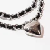 Gargantilla de doble cadena, una de eslabones plateados y otra de cordón trenzado negro, con dije de corazón plateado.