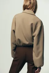 Chaqueta bomber negra con cuello solapa, manga larga abullonada y puños elásticos. Presenta falsos bolsillos de doble vivo en la parte delantera y cierre frontal con cremallera.