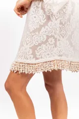 Vestido camisero corto de encaje blanco con diseño ornamental, escote en V con cordón ajustable y ojales metálicos, mangas 3/4 acampanadas y ruedo con detalle de flecos de caracoles tejidos a mano.