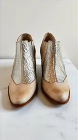 Botas cortas de cuero color natural con inserciones de cuero plateado con textura de cocodrilo en el empeine, paneles laterales elásticos y detalles pintados a mano en el talón. Cuenta con taco de madera de 5 cm.