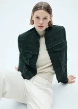 Chaqueta de tweed en color verde oscuro, con diseño corto y recto. Presenta cuello alto, cierre frontal con botones, mangas largas y dos bolsillos de solapa con botón en el frente. Se caracteriza por sus terminaciones deshilachadas y costuras decorativas.