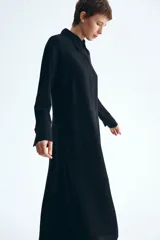 Vestido midi negro con cuello camisero y tapeta con botones ocultos, de mangas largas raglán con puños anchos y costura fruncida en la cadera con falda de vuelo leve.