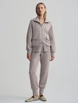 Campera deportiva con cierre frontal, cuello alto tipo chimenea y mangas raglán. Es de corte holgado y presenta bolsillos de parche delanteros. El color es un tono neutro entre gris y rosado (taupe).