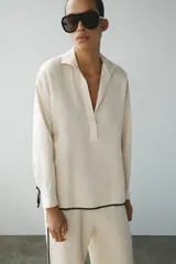 Camisa de lino color crudo con cuello solapa, manga larga y bajo asimétrico. Presenta detalles de ribetes negros a contraste y aberturas laterales con tiras para ajustar.
