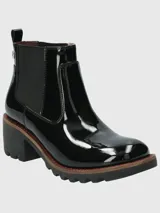 Bota tipo Chelsea de caña corta en charol negro, con paneles laterales elásticos, suela track robusta y taco cuadrado. Presenta un detalle de costura en color rojo en el borde de la suela.
