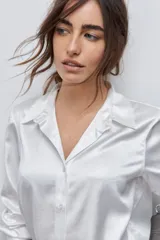 Camisa blanca de calce relajado con acabado satinado y brillo sutil, cuello clásico, cierre frontal con botones y mangas largas con puños.