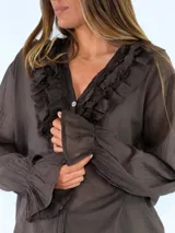 Blusa de manga larga color marrón, con cierre frontal de botones, escote en V y detalles de volados fruncidos alrededor del cuello y los puños elásticos.