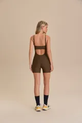 Jumpsuit corto deportivo color marrón con breteles finos ajustables, escote con abertura y bolsillos laterales. Confeccionado con poliamida reciclada y elastano, ofrece protección UV 50+ y tecnología Easy Care.