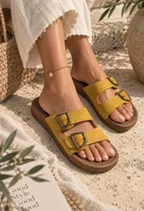 Sandalias planas de mujer con doble tira en tono mostaza de acabado gamuzado, sujetas por dos hebillas metálicas de estilo rústico y base marrón.