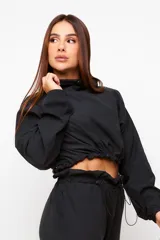Buzo cropped negro de estilo urbano, con cuello alto ajustable mediante cordón, mangas largas con puños elásticos y cintura con ajuste regulable.