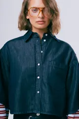 Camisa de denim azul de corte cropped, manga corta ancha, cuello camisero, cierre frontal de botones y un bolsillo aplicado en el pecho.